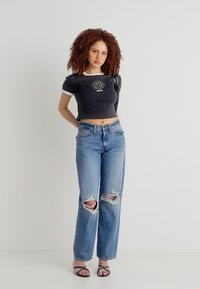 Νεαρή γυναίκα με σγουρά μαλλιά που φοράει μαύρο κοντό μπλουζάκι με γραφικό ήλιο, σκισμένο μπλε τζιν και μαύρα σανδάλια με λουράκια, στέκεται δίπλα σε λευκό τοίχο.