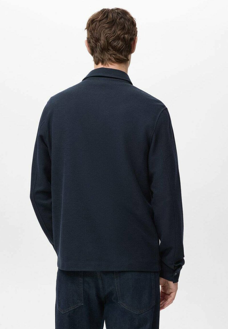 Polo à manches longues bleu marine avec une finition en maille texturée, doté d'un col et de détails à boutons, associé à un pantalon en denim foncé.