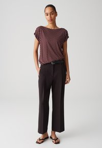 Burgunderfarbener Kurzarm-Top mit geometrischem Muster, kombiniert mit schwarzen hoch taillierten Hosen. Das Model trägt schwarze flache Sandalen.