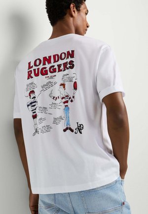 Homme portant un t-shirt blanc avec le texte rouge « LONDON RUGGERS » et un dessin de joueurs de rugby dessin animé dans le dos, associé à un jean bleu clair.