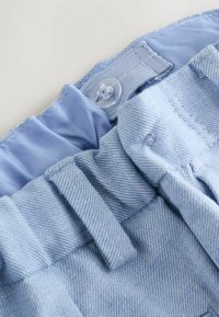 Svetlo modra denim tkanina z pasom, ki ima zapiranje na gumb in elastični notranji sloj, prikazuje mehko teksturo in subtilen črtast vzorec.