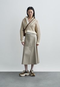 Beige, strikket cropped sweater med tekstureret mønster, lagdelt over en cremefarvet top, kombineret med en beige A-formet nederdel og chunky sneakers.