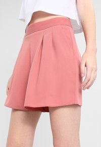 Skládané šortky z měkké růžové látky s hladkou texturou a pasovým lemem. Design má rozšířený střih, který sahá do poloviny stehen.