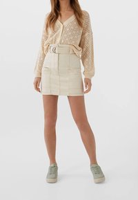 Beige stickad cardigan med lös passform och V-ringning, kombinerad med en hög midja denimkjol och ljusgröna sneakers.