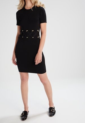 Femme portant une robe noire cintrée à manches courtes, une large ceinture tressée et des chaussures noires sans lacets avec des boucles dorées.