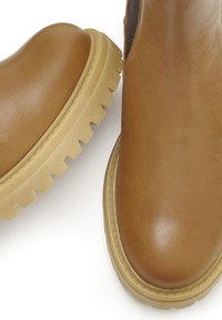 Tan leren enkelboots met een glanzende afwerking, voorzien van een chunky beige rubberen zool met diepe groeven voor grip.