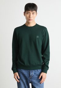 Groene sweatshirt met een ronde hals en ribgebreide manchetten, voorzien van een kleine vierkante logopatch op de linkerkant van de borst; gecombineerd met blauwe jeans.