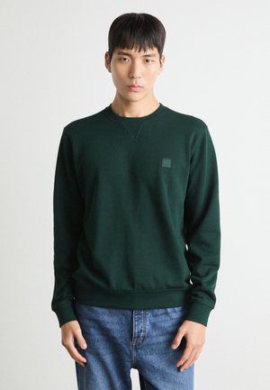 Green sweatshirt with a round neckline and ribbed cuffs, featuring a small square logo patch on the left chest; paired with blue jeans.