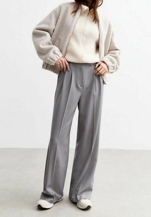Person trägt eine beige Jacke zum Reißverschluss, einen cremefarbenen gerippten Pullover, weite graue Hosen und weiße Sneakers und steht vor einem schlichten Hintergrund.