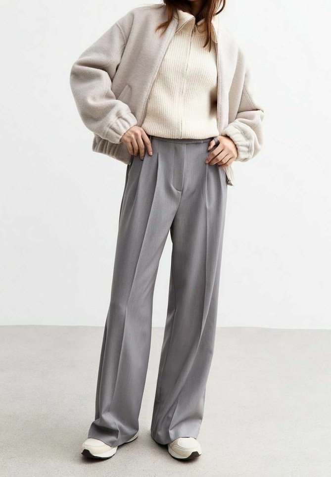 Personne portant une veste beige à fermeture éclair, un pull côtelé crème, un pantalon large gris et des baskets blanches, debout devant un fond uni.