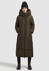 Žena v dlhom tmavo olivovom prešívanom kabáte s vysokým golierom, čiernej čiapke, čiernych nohaviciach a čiernych čižmách, stojí so zarovenými rukami vo vreckách.