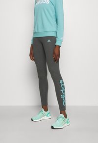 Ljusblå sweatshirt med vit logotyp, i kombination med mörkgrå leggings som har blå branding. Modellen bär mintgröna löparskor.