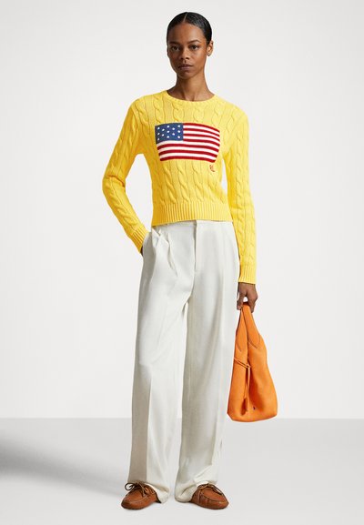 Rumena pulover s pletenim vzorcem in motivom ameriške zastave, kombiniran z belimi širokimi hlačami in oranžno torbico.