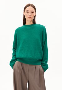 Frau mit glattem braunem Haar, die einen übergroßen grünen Pullover und lockere beigefarbene Hosen trägt, mit Händen in den Taschen vor einem weißen Hintergrund.