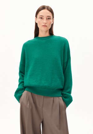 Woman with straight brown hair wearing an oversized green sweater and loose beige pants with hands in pockets on a white background.