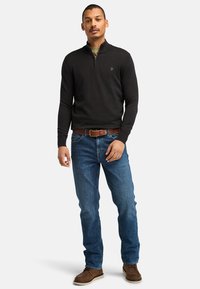 Sort lynlås-trøje med lange ærmer, blå denim jeans, brun læderbælte og brune støvler, med et logo på trøjen.