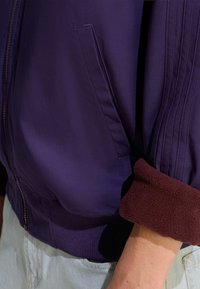 Violetti vetoketjullinen takki, jossa on ribbireunus ja hihansuut. Hihansuissa on burgundin väristä fleece-yksityiskohtaa. Sivutasku näkyy sauman lähellä. Tasainen pintatekstuuri.