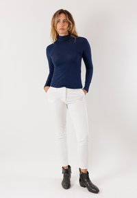 Maglietta a maniche lunghe blu navy con collo alto, design aderente, abbinata a pantaloni bianchi slim-fit. Stivaletti neri alla caviglia con finitura in pelle testurizzata.