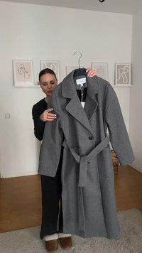 Ανοιχτό γκρι μάλλινο παλτό με μεγάλο γιακά, κουμπωτό κλείσιμο και ζώνη από ύφασμα; διαθέτει μακριά μανίκια και ίσια γραμμή.