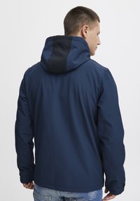 Tumši zila jaka ar kapuci, kurai ir kontrastējoša melna paneļa aizmugurē, gluda tekstūra un minimālistisks dizains.