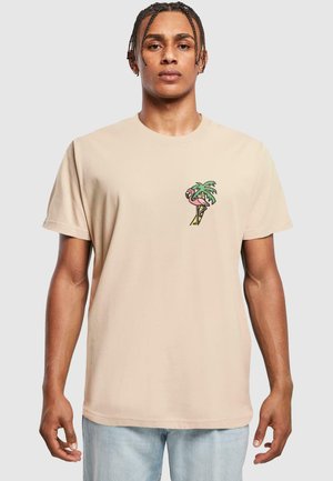 Camiseta de algodón beige con cuello redondo y mangas cortas, que presenta un gráfico colorido de una palmera y un flamenco en el lado izquierdo del pecho.