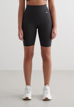 Vrouw draagt zwarte, hoogtallige geribbelde biker shorts en witte sneakers, staand tegen een effen lichte achtergrond.