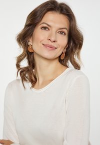 Hellbeige Langarmoberteil mit rundem Ausschnitt. Das Modell trägt große, runde, halbtransparente Ohrringe in Pfirsich- und Brauntönen.