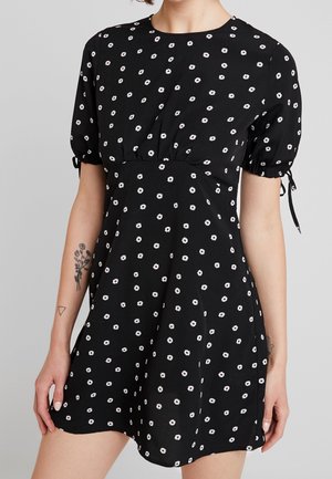 Zwarte korte mouwen jurk met witte madeliefjesprint, getailleerde taille en gebonden mouwdetails, gedragen door een persoon met bloemtattoo's op de linker onderarm en hand.