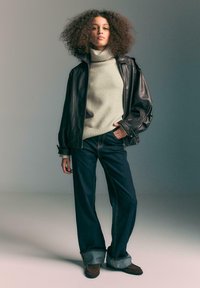 Giacca di pelle nera, maglione a collo forte crema, jeans a gamba larga in denim scuro con orli risvoltati, scarpe marroni e capelli ricci naturali.