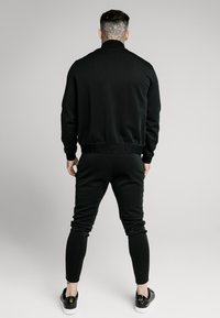 Chándal atlético negro con chaqueta con cremallera y cuello alto, puños acanalados y pantalones ajustados a juego. Tejido suave y diseño minimalista.