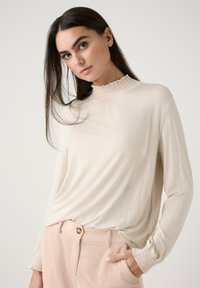 Langarm-blaues Bluse in Beige mit einem hohen, gerafften Kragen und strukturierten Manschetten, kombiniert mit hellrosanen Ripp-Hosen mit einem goldenen Knopf.