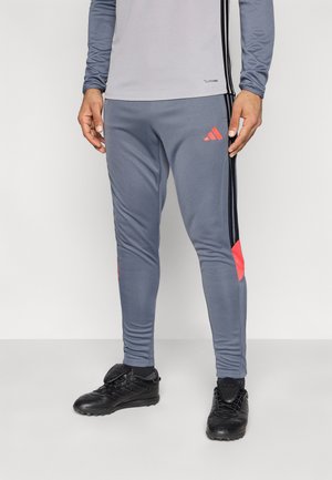 Pantalones deportivos grises con franjas laterales negras y detalles en rojo. Hechos de tela elástica, con un diseño ajustado y cintura elástica.