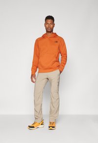 Felpa arancione con tasca frontale, pantaloni cargo beige e scarpe da ginnastica gialle e nere. Modello in piedi su uno sfondo semplice.