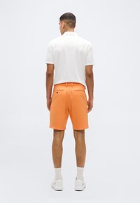 Άνδρας που κοιτάζει προς τα πίσω φορώντας λευκό πόλο πουκάμισο, πορτοκαλί σορτς με πίσω τσέπες, λευκές κάλτσες και λευκά αθλητικά παπούτσια σε απλό φόντο.