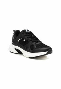 Zapatilla deportiva negra con superposiciones de malla y sintético, con acentos blancos, punta redondeada y una gruesa entresuela blanca. Cordones negros y logo presentes.