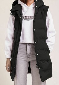 Czarna pikowana kamizelka z wysokim kołnierzem, zamkiem z przodu i zatrzaskami. Noszona na białej bluzie z kapturem i uzupełniona jasnoszarymi dżinsami.