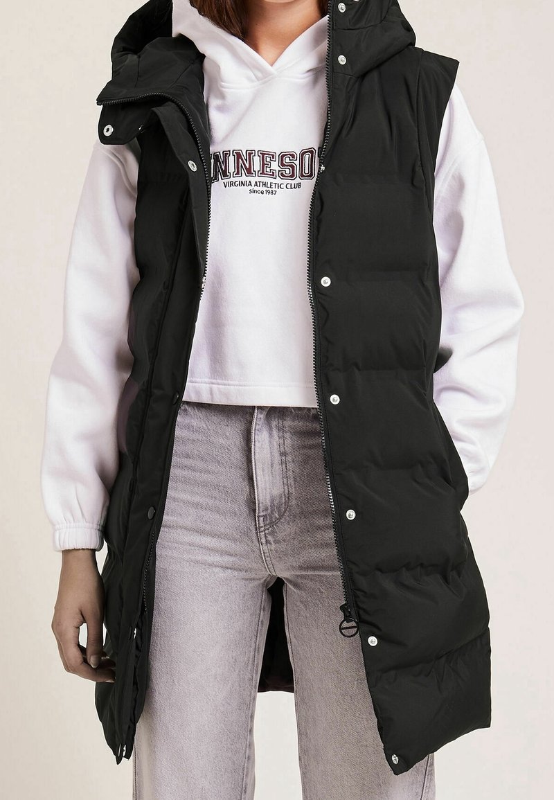 Czarna pikowana kamizelka z wysokim kołnierzem, zamkiem z przodu i zatrzaskami. Noszona na białej bluzie z kapturem i uzupełniona jasnoszarymi dżinsami.