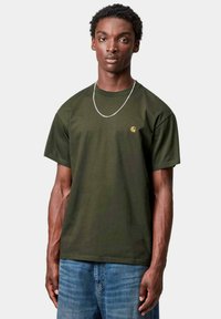 T-shirt in cotone verde scuro con maniche corte, con un piccolo logo giallo sul lato sinistro del petto. Indossato con jeans in denim blu.