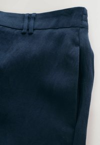 Zona taliei pantalonilor bleumarin cu găici pentru curea și buzunar lateral parțial vizibil pe un fundal deschis.