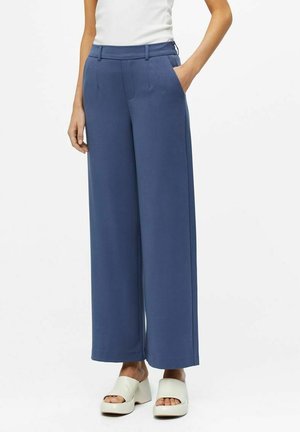 Person wearing high-waisted, wide-leg blue trousers with side pockets, paired with white open-toe platform sandals and a white sleeveless top.