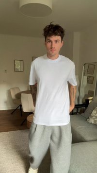 Tricou alb cu mânecă scurtă asociat cu pantaloni de trening gri, prezentat într-un decor de living cu mobilier și decorațiuni neutre.