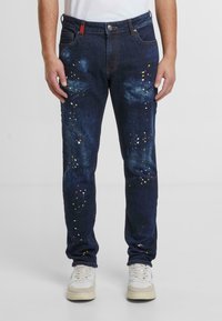 Tmavě modré džíny s rovnými nohavicemi, zdobené skvrnami od barvy v červené, žluté a bílé barvě, s designem pěti kapes a poutky na opasek.