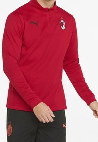 Punainen pitkähihainen urheilu-paita, jossa on vetoketjulla varustettu kaulus. Paidassa on musta Puma-logo ja AC Milan -embleemi rinnassa. Sileä kangaspinta.