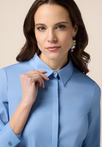 Cămașă lungă cu mânecă, de culoare albastru deschis, cu guler ascuțit, detaliu cu nasturi la gât și textură netedă. Modelul poartă cercei mari.
