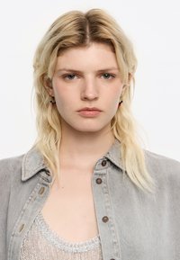 Hellgraue Jeansjacke mit braunen Knöpfen, über einem silbernen, metallischen Oberteil getragen. Das Model hat langes, glattes, blondes Haar und trägt rote und schwarze Ohrringe.