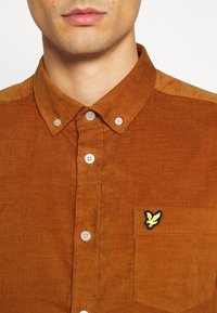 Camisa de pana marrón con cuello abotonado, que presenta botones blancos y un logo de un pájaro amarillo en el bolsillo del pecho izquierdo.