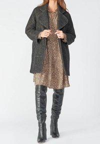 Cappotto nero texturizzato con ampi risvolti sovrapposto a un vestito con stampa leopardo. Stivali in pelle nera aderenti, alti fino al ginocchio, completano il look.