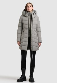 Vadderad grå parka-jacka med huva, med vertikal quiltad design, dragkedjestängning och svarta accenter. Bärs över svarta tights och stövlar.