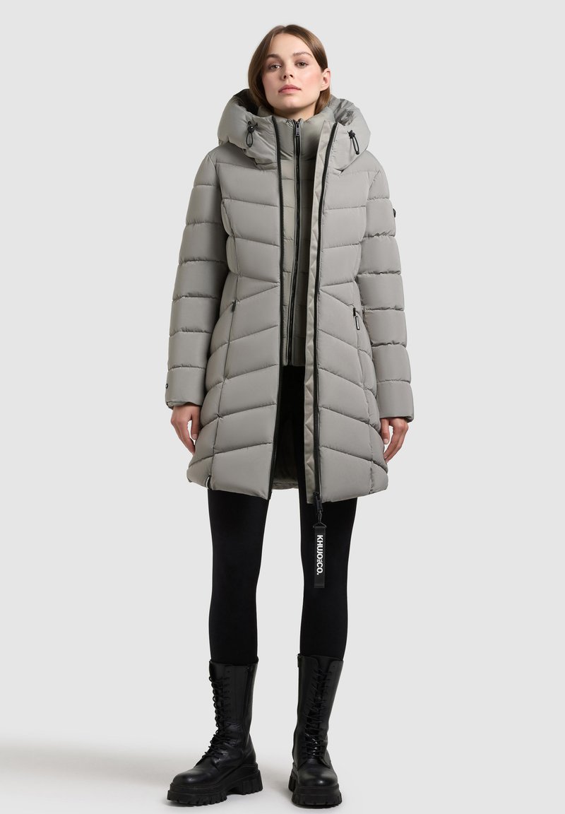 Geacă parka căptușită de culoare gri, cu glugă, având un design vertical matlasat, închideri cu fermoar și accente negre. Purtați-o peste colanți negri și cizme.