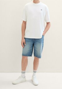 Valkoinen lyhythihainen t-paita, jossa on minimaalinen logo, yhdistettynä vaaleansinisiin farkkushortseihin. Asusteena valkoiset tennarit ja raidalliset sukat.